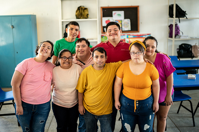 Eight teenagers with disabilities happily standing close together, wearing different colored shirts and smiling. They are in a recreation room. 